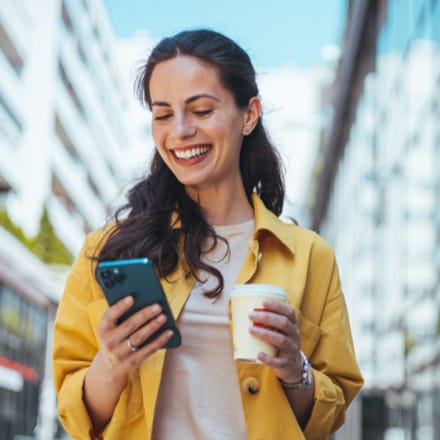 A smiling woman holding a cup of coffee and looking at her mobile device.