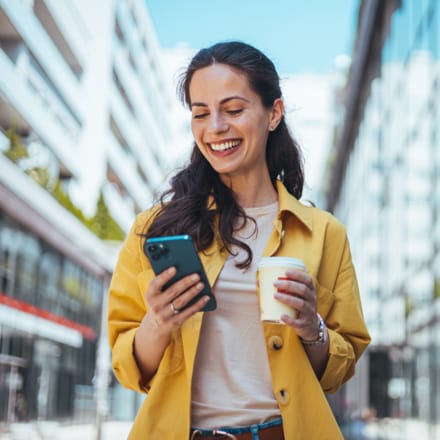A smiling woman holding a cup of coffee and looking at her mobile device.