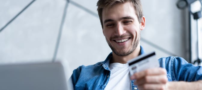 A young man holding a debi card while looking at a laptop.