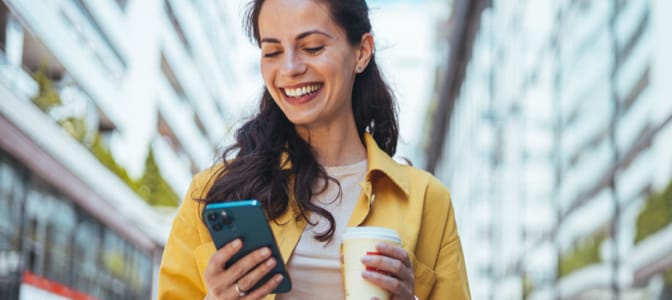 A smiling woman holding a cup of coffee and looking at her mobile device.