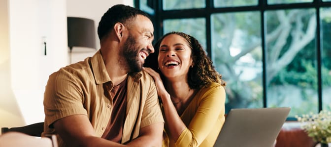 A couple smiling at each other sitting in front of a laptop.